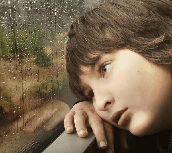 A young boy rests his head on a window sill staring into space through a window on a rainy day.
